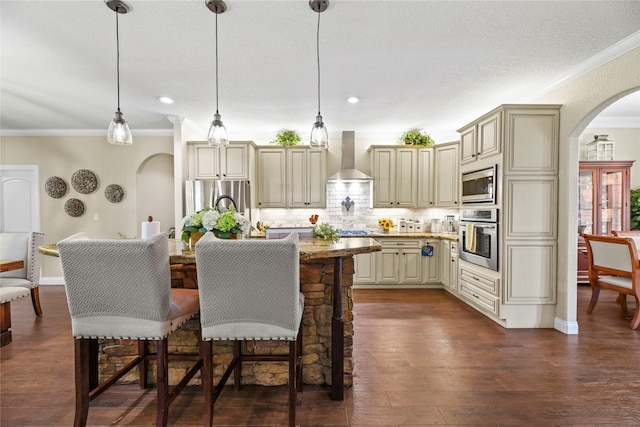 kitchen featuring appliances with stainless steel finishes, cream cabinets, arched walkways, ornamental molding, and wall chimney exhaust hood