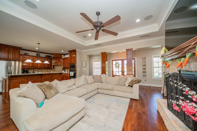 living room featuring dark wood finished floors, a stone fireplace, a ceiling fan, decorative columns, and a tray ceiling