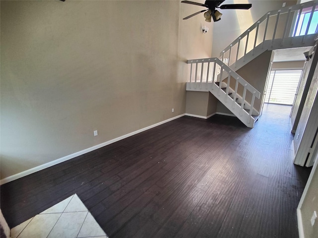 unfurnished living room with a ceiling fan, a towering ceiling, wood finished floors, baseboards, and stairs