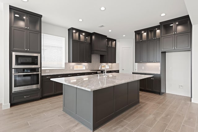 kitchen featuring decorative backsplash, a kitchen island with sink, built in microwave, light wood-style flooring, and oven