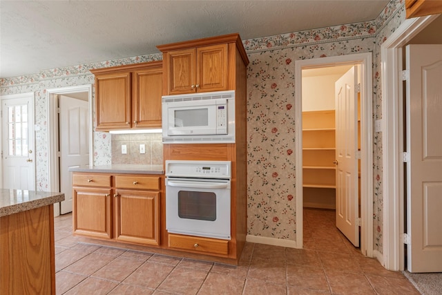 kitchen with brown cabinetry, light tile patterned floors, baseboards, wallpapered walls, and white appliances