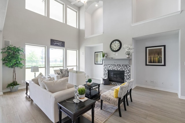 living area featuring ceiling fan, light wood finished floors, a tiled fireplace, baseboards, and a towering ceiling