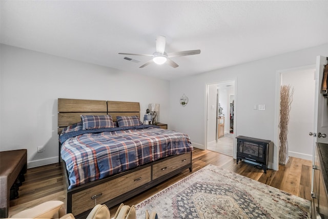bedroom featuring a wood stove, a ceiling fan, baseboards, wood finished floors, and visible vents