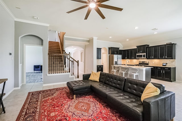 living room with crown molding, light tile patterned flooring, arched walkways, a ceiling fan, and stairway