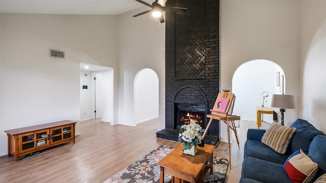 living room with ceiling fan, visible vents, a brick fireplace, high vaulted ceiling, and light wood finished floors
