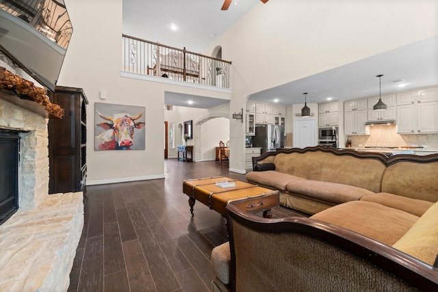 living room with ceiling fan, arched walkways, a stone fireplace, baseboards, and dark wood-style floors