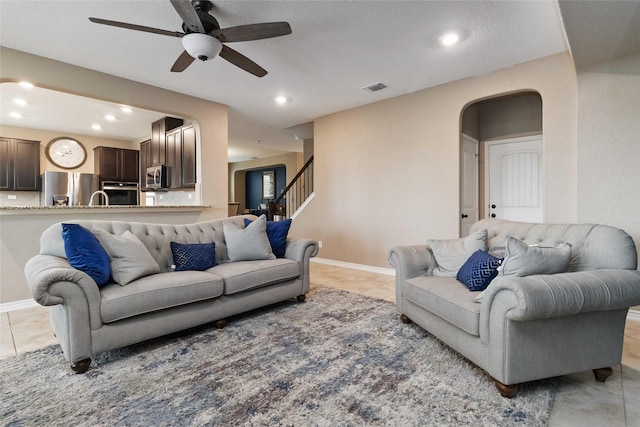 living room with light tile patterned flooring, ceiling fan, arched walkways, visible vents, and stairway