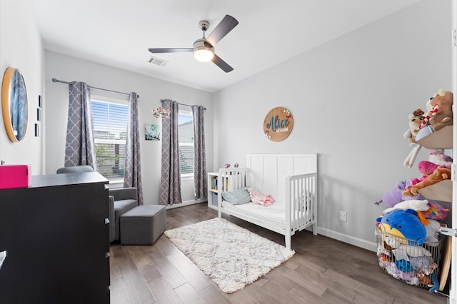 bedroom featuring ceiling fan, visible vents, wood finished floors, a crib, and baseboards