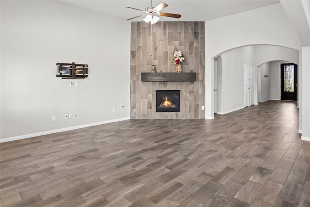 unfurnished living room with ceiling fan, arched walkways, a tiled fireplace, baseboards, and wood finished floors