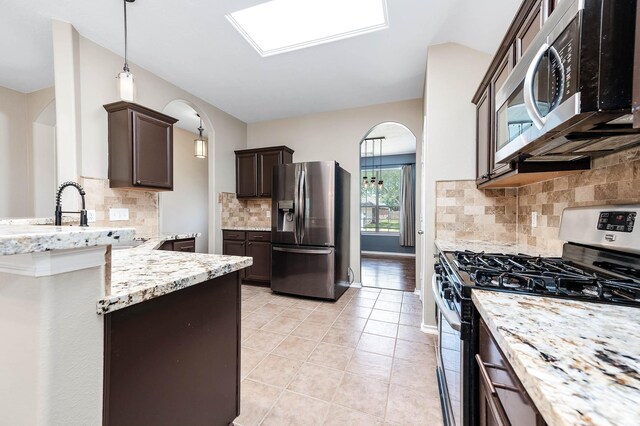 kitchen featuring appliances with stainless steel finishes, arched walkways, dark brown cabinetry, light stone counters, and tasteful backsplash