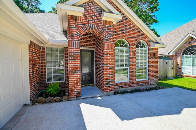 entrance to property featuring fence, brick siding, a shingled roof, an attached garage, and driveway
