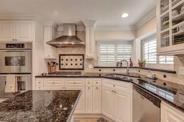 kitchen with white cabinetry, a sink, dark stone countertops, stainless steel appliances, and wall chimney exhaust hood