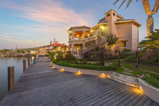 back of property at dusk featuring fence, a balcony, a water view, stairs, and stucco siding