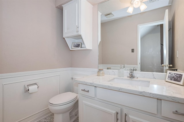 half bath featuring wainscoting, a decorative wall, vanity, toilet, and visible vents