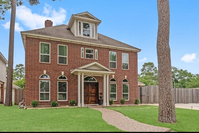 colonial home with a chimney, a front yard, brick siding, fence, and a shingled roof