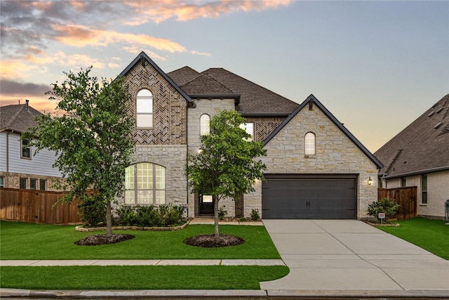 french country inspired facade with a lawn, fence, concrete driveway, and stone siding
