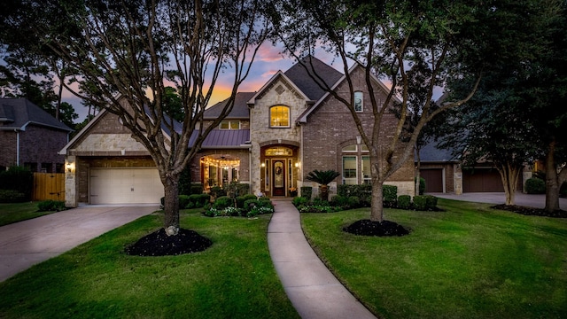 french provincial home featuring an attached garage, stone siding, a standing seam roof, concrete driveway, and a lawn