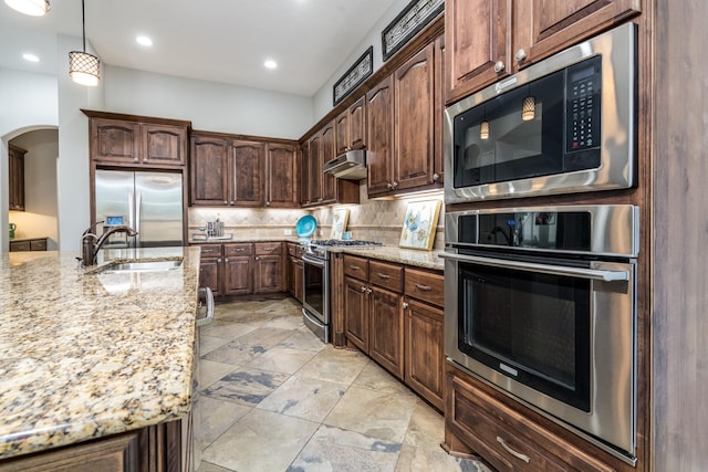 kitchen featuring appliances with stainless steel finishes, light stone counters, a sink, arched walkways, and decorative backsplash
