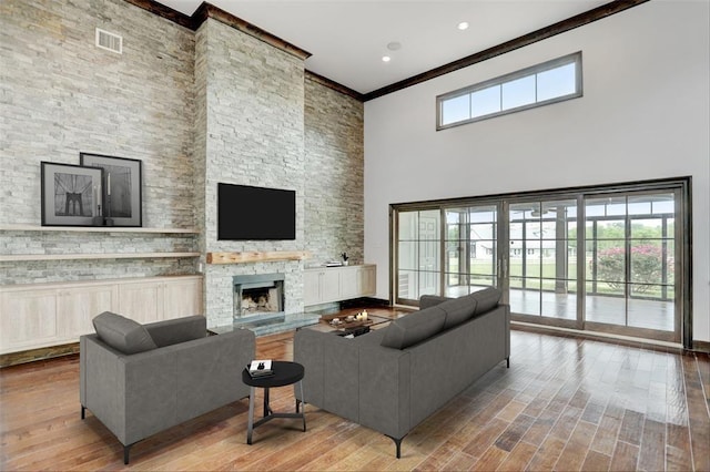 living room featuring a stone fireplace, visible vents, a towering ceiling, wood finished floors, and ornamental molding