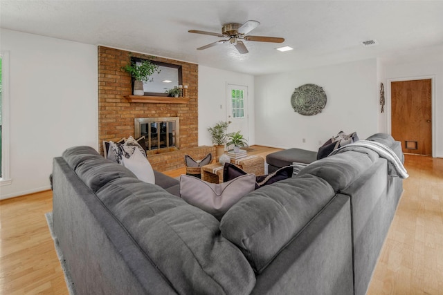 living room with a brick fireplace, a ceiling fan, baseboards, light wood-type flooring, and visible vents