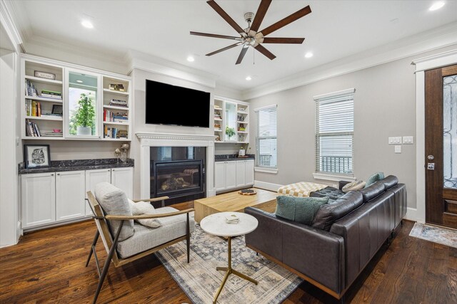living area featuring dark wood-style flooring, a glass covered fireplace, a ceiling fan, baseboards, and ornamental molding