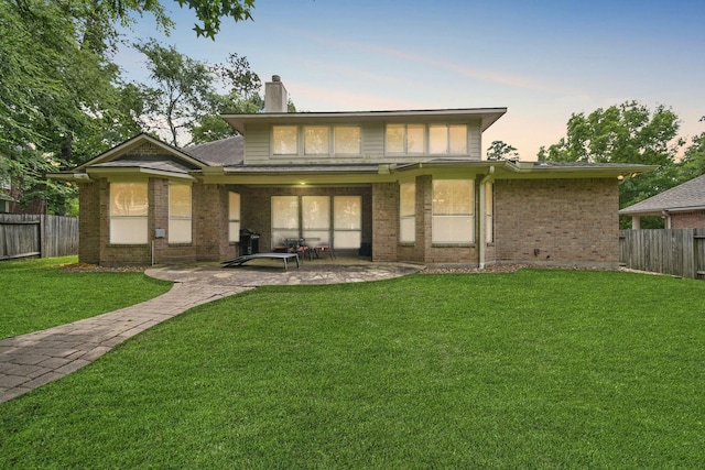 back of house at dusk with a fenced backyard, a chimney, brick siding, a patio area, and a yard