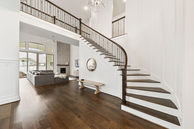 staircase featuring a towering ceiling, a large fireplace, wood finished floors, a chandelier, and a decorative wall