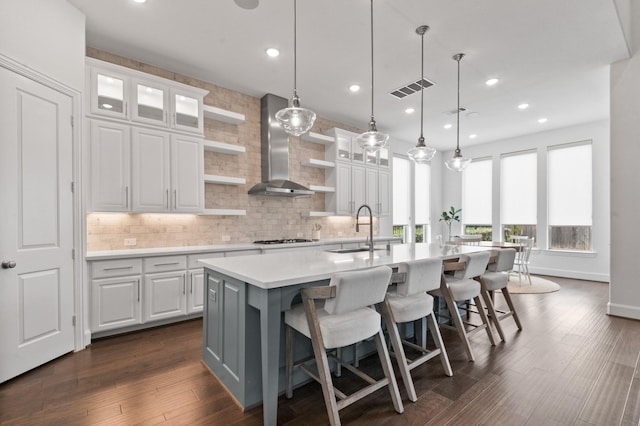 kitchen featuring wall chimney range hood, a kitchen island with sink, open shelves, a sink, and light countertops