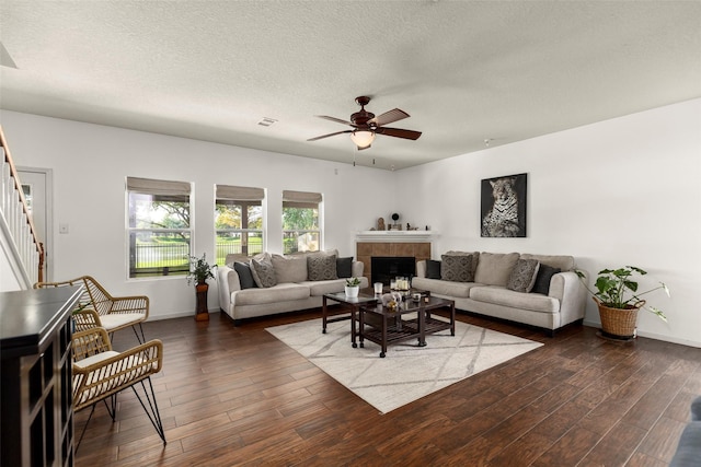 living room with a ceiling fan, a textured ceiling, a tile fireplace, stairway, and dark wood-style floors