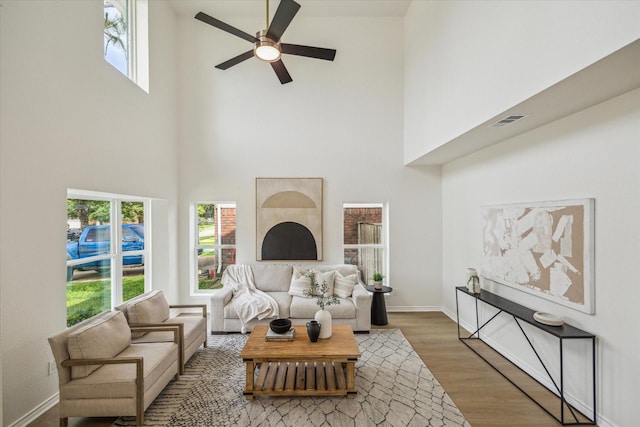 living room with a ceiling fan, a healthy amount of sunlight, wood finished floors, a towering ceiling, and visible vents
