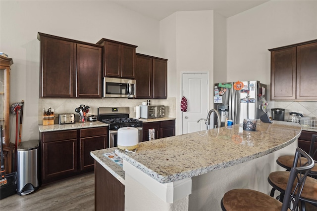 kitchen with appliances with stainless steel finishes, tasteful backsplash, light stone countertops, dark brown cabinets, and a breakfast bar
