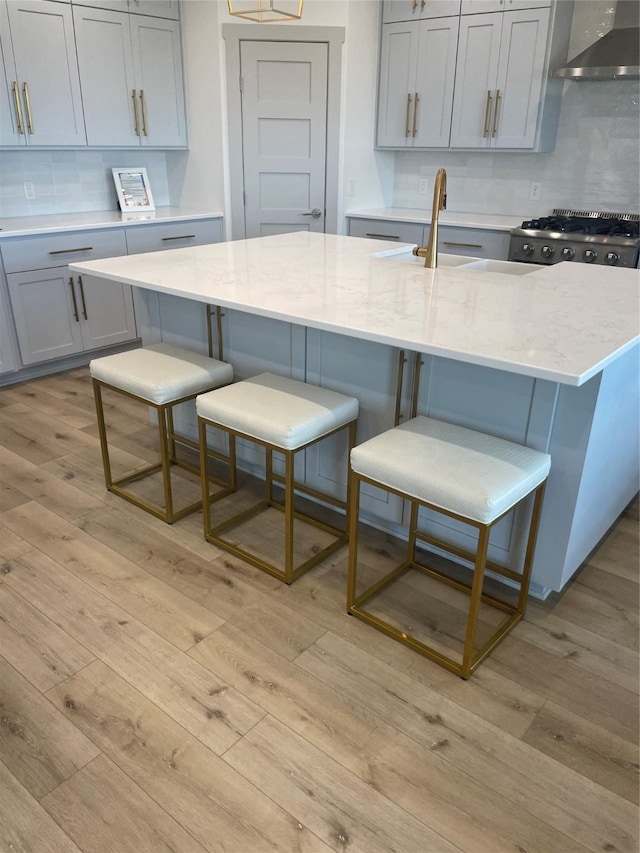 kitchen with wall chimney range hood, gray cabinetry, light stone counters, a breakfast bar, and light wood-style flooring