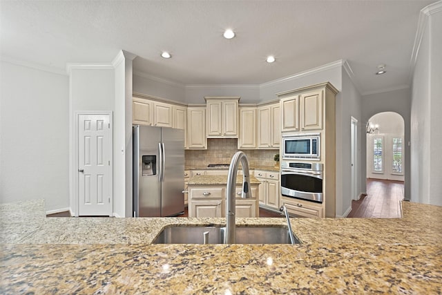 kitchen with decorative backsplash, arched walkways, cream cabinets, a sink, and appliances with stainless steel finishes