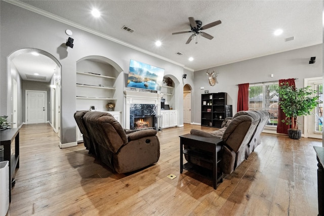 living room featuring crown molding, ceiling fan, built in shelves, a tile fireplace, and arched walkways
