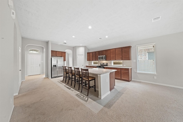 kitchen featuring a kitchen island with sink, baseboards, light colored carpet, arched walkways, and stainless steel appliances