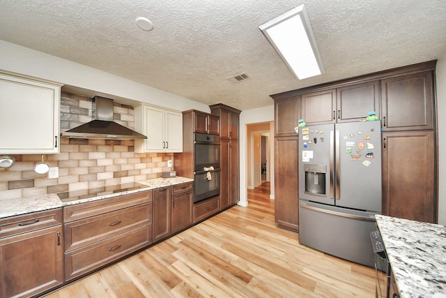 kitchen with a textured ceiling, visible vents, black appliances, light wood finished floors, and wall chimney exhaust hood