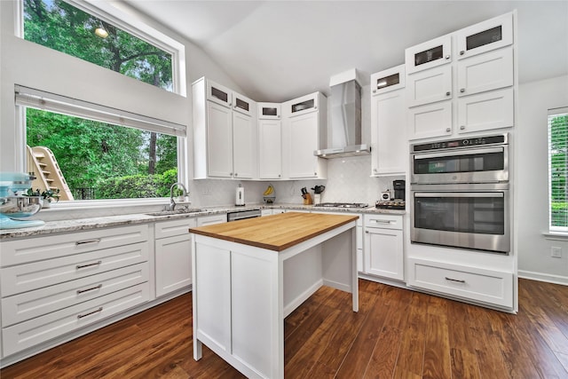 kitchen with wall chimney range hood, backsplash, lofted ceiling, wood counters, and stainless steel appliances