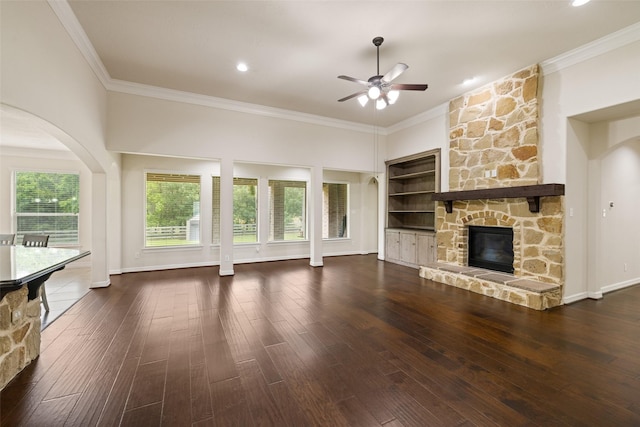 unfurnished living room featuring dark wood-style flooring, arched walkways, a stone fireplace, a ceiling fan, and ornamental molding