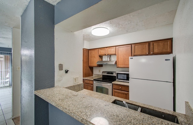kitchen featuring wall chimney range hood, brown cabinets, a textured wall, stainless steel appliances, and light tile patterned flooring