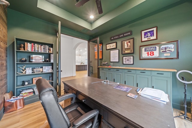 home office featuring arched walkways, a raised ceiling, a ceiling fan, and wood finished floors