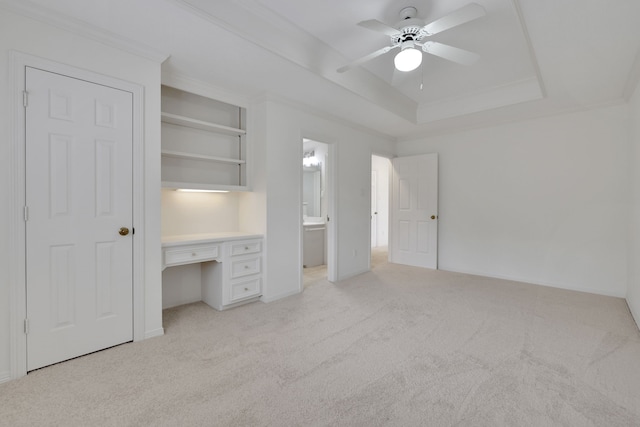 unfurnished bedroom with ceiling fan, built in desk, a raised ceiling, light carpet, and ensuite bath