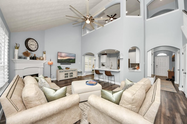 living area with dark wood-style flooring, a fireplace, arched walkways, high vaulted ceiling, and a ceiling fan