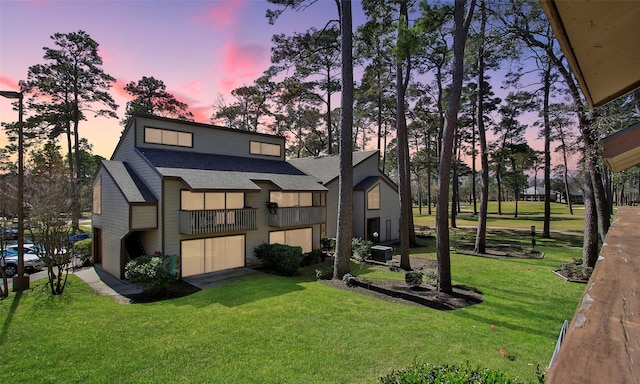 back of property at dusk featuring roof with shingles, an attached garage, a lawn, and a balcony