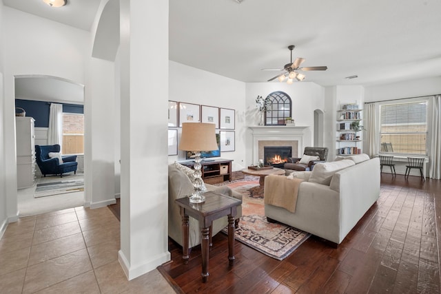 living area with ceiling fan, arched walkways, a glass covered fireplace, baseboards, and wood finished floors