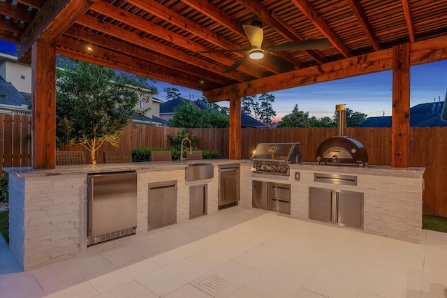 patio terrace at dusk featuring a fenced backyard, a sink, a grill, and an outdoor kitchen