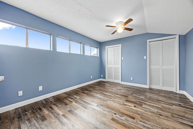 unfurnished bedroom featuring a textured ceiling, two closets, baseboards, wood finished floors, and vaulted ceiling