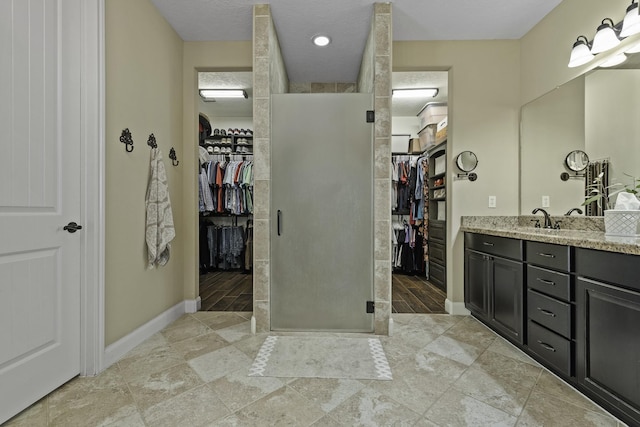 full bathroom with a walk in closet, a stall shower, vanity, baseboards, and stone tile floors