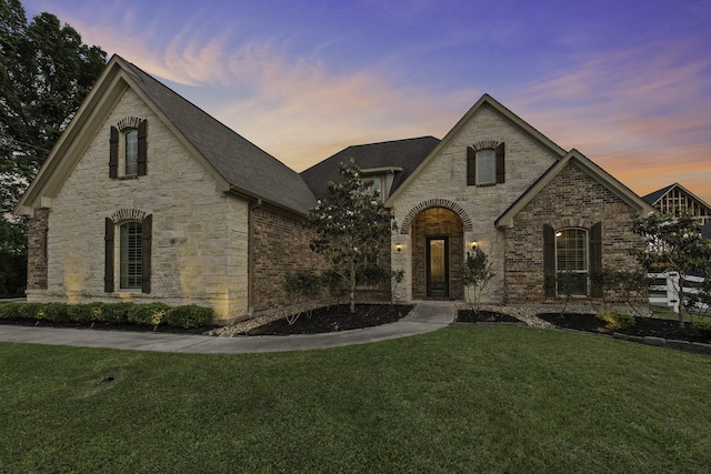 french country home featuring roof with shingles, stone siding, brick siding, and a front yard