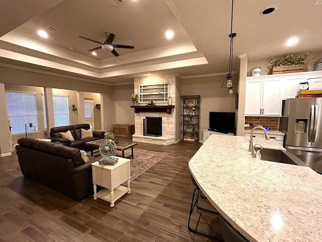 living area featuring a raised ceiling, a fireplace, ornamental molding, a ceiling fan, and dark wood-style floors