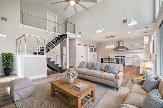 living room featuring ceiling fan, visible vents, a barn door, stairway, and high vaulted ceiling